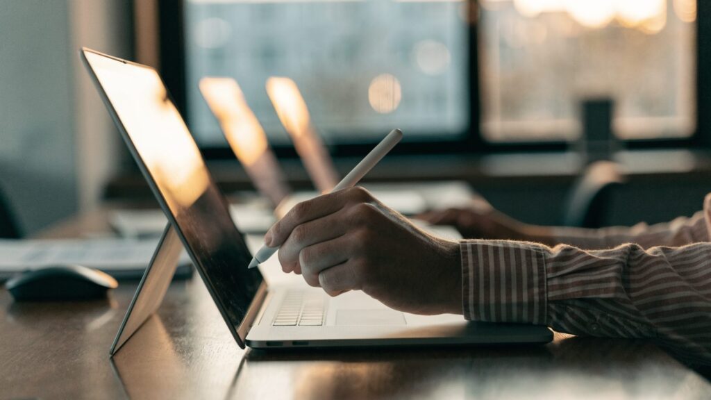 A hand drawing with a stylus on a 2-in-1 silver laptop in tablet mode on an office desk
