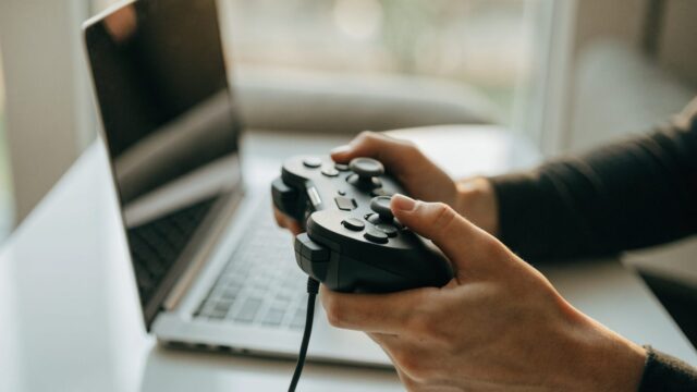 Hands holding a game controller in front of a silver laptop on a white desk