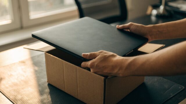 Hands lifting a brand-new graphite black laptop out of its plain cardboard box on a dark desk mat