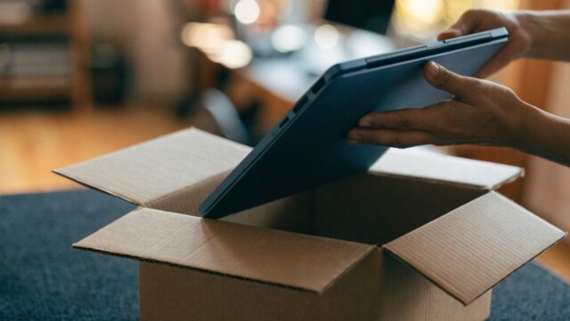 Hands lifting a brand-new dark blue laptop out of its plain cardboard box on a dark desk mat