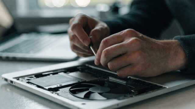 A technician's hands holding a small screwdriver over an opened silver laptop with internal components visible