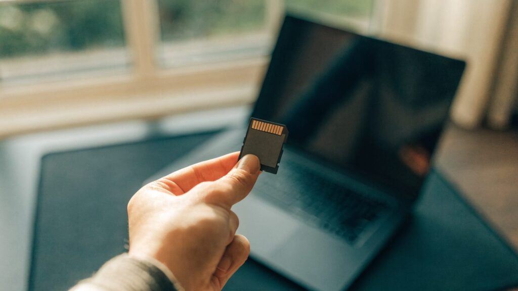 An SD card held between two fingers in front of a graphite black laptop on a dark desk mat
