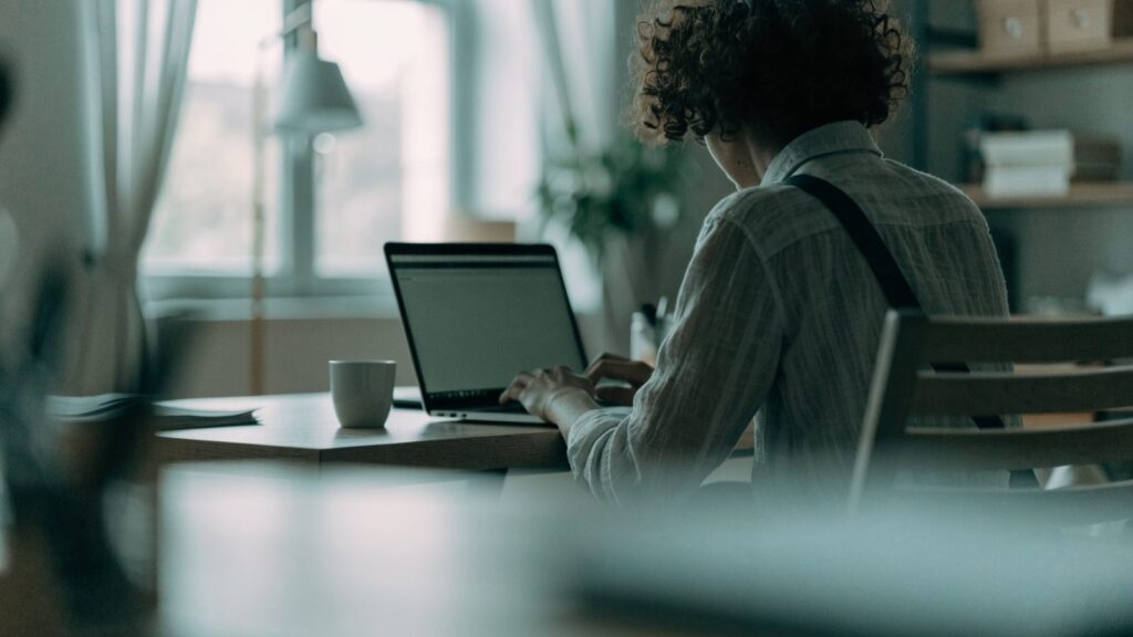 A person seen from behind working on a silver laptop in a bright home office