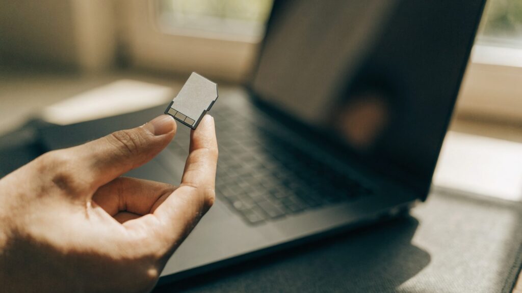 An SD card held between two fingers in front of a graphite black laptop on a dark desk mat
