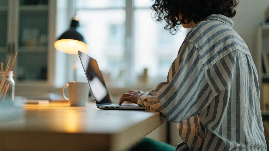 A person seen from behind working on a silver laptop in a bright home office