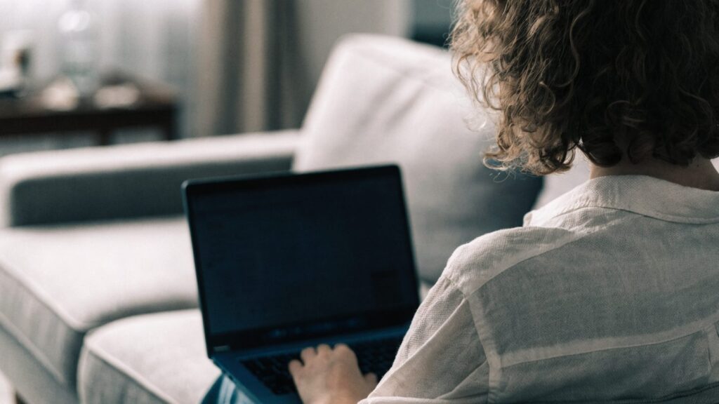 A person seen from behind using a dark blue laptop on a sofa