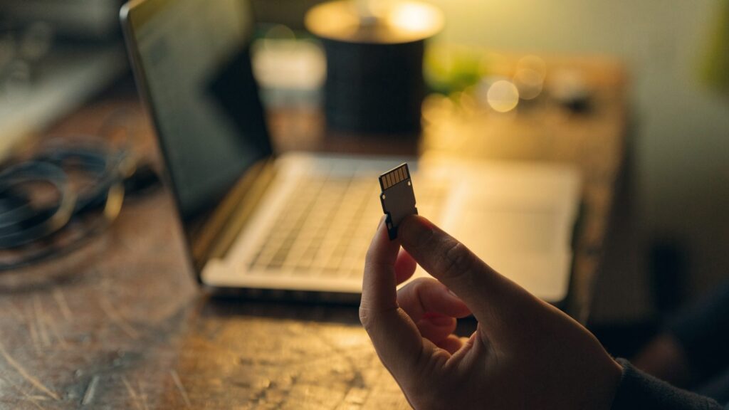 An SD card held between two fingers in front of a silver laptop on a workbench
