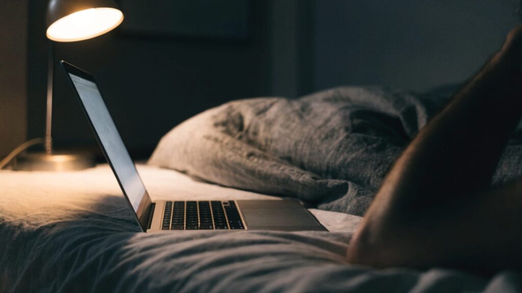 A matte grey laptop glowing softly on a bed in a dark room