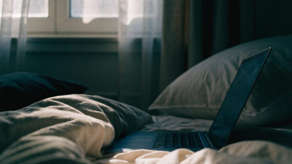 A dark blue laptop glowing softly on a bed in a dark room