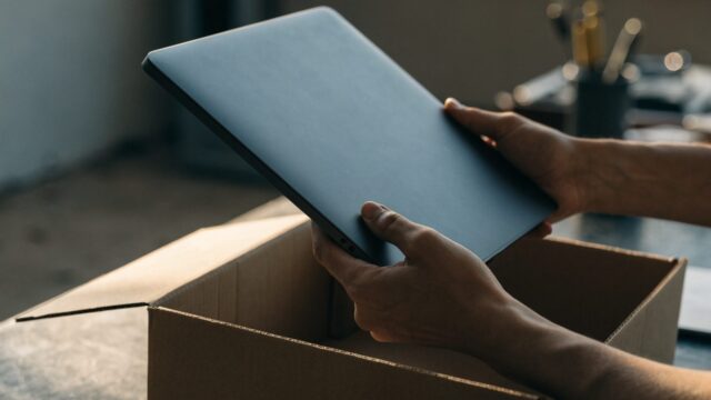 Hands lifting a brand-new graphite black laptop out of its plain cardboard box on a workbench