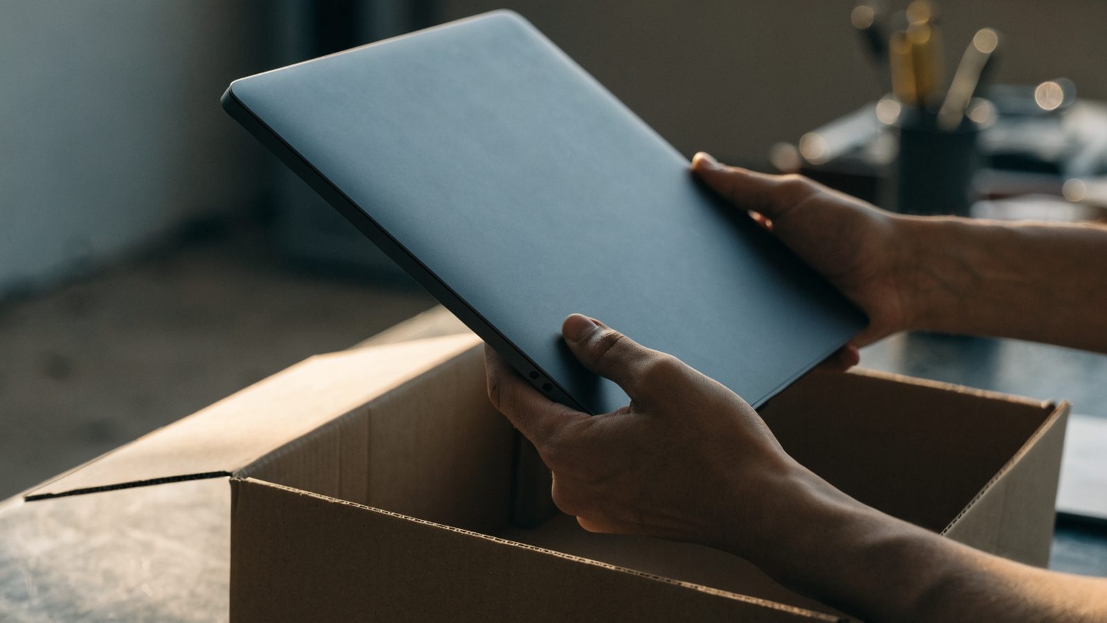 Hands lifting a brand-new graphite black laptop out of its plain cardboard box on a workbench