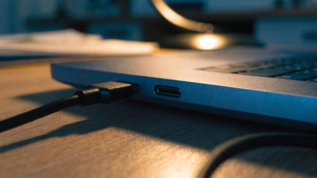 Close-up of a USB-C charger cable plugged into the side port of a dark blue laptop on a wooden desk