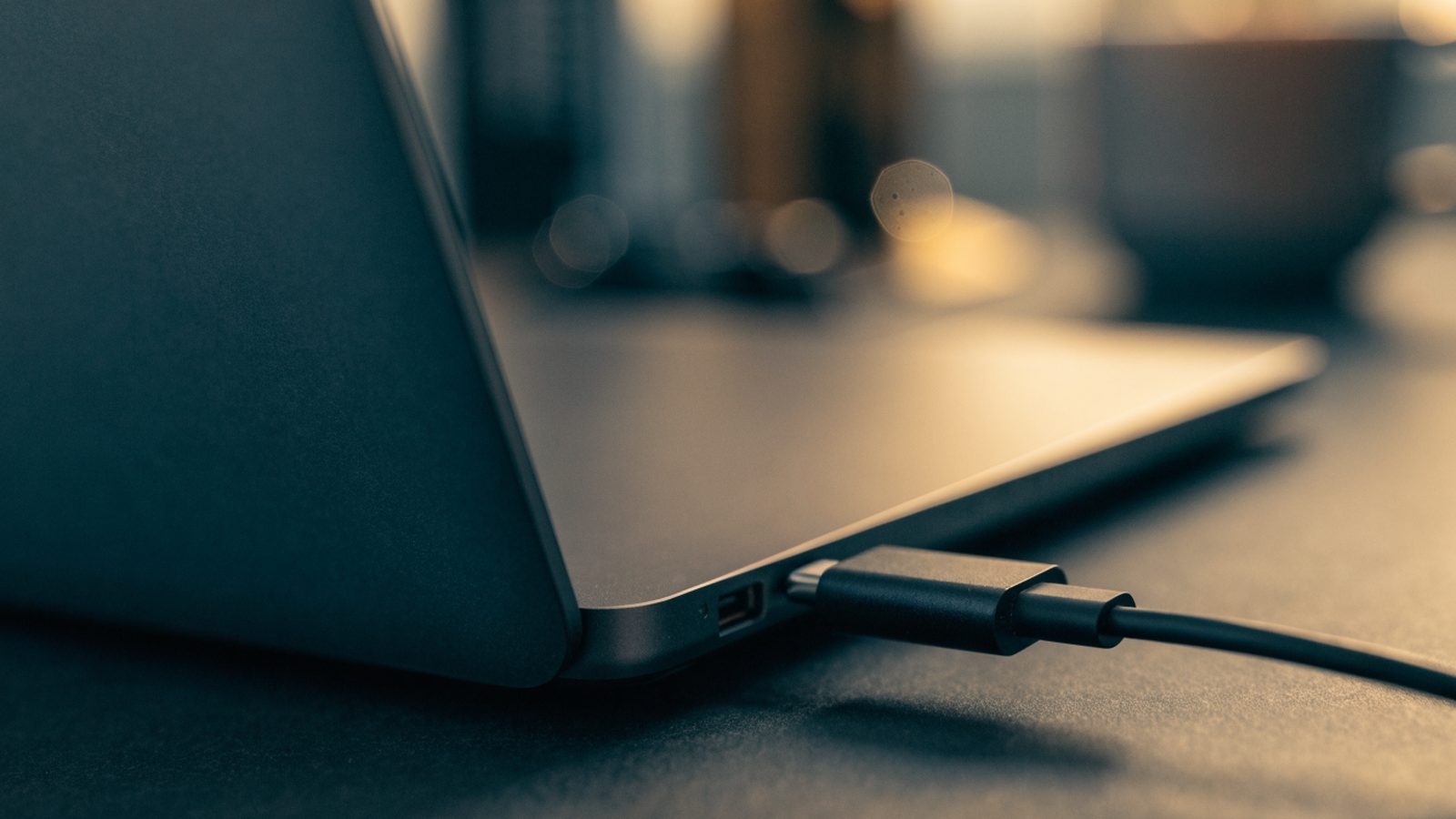 Close-up of a USB-C charger cable plugged into the side port of a graphite black laptop on a dark desk mat
