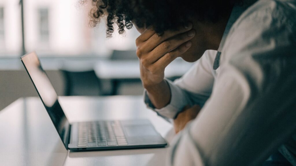A person resting their forehead on one hand in front of a graphite black laptop on a white desk
