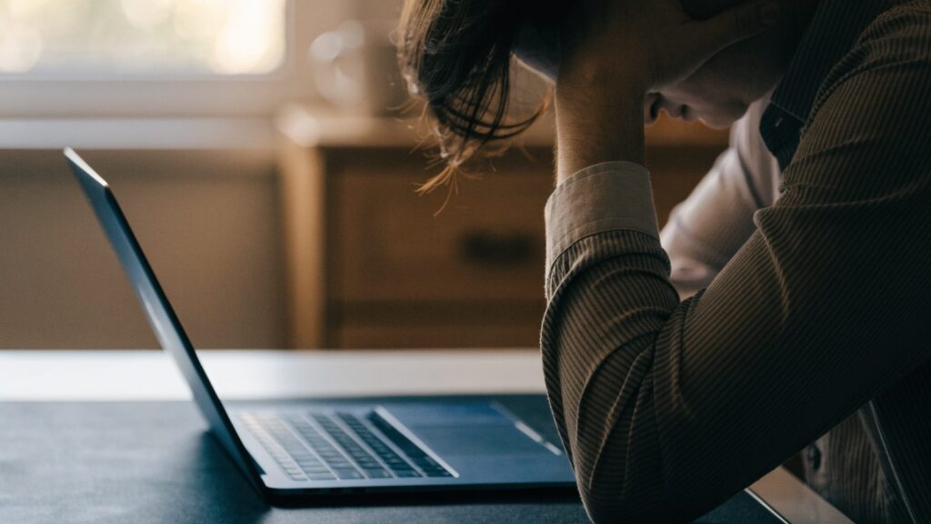 A person resting their forehead on one hand in front of a dark blue laptop on a dark desk mat