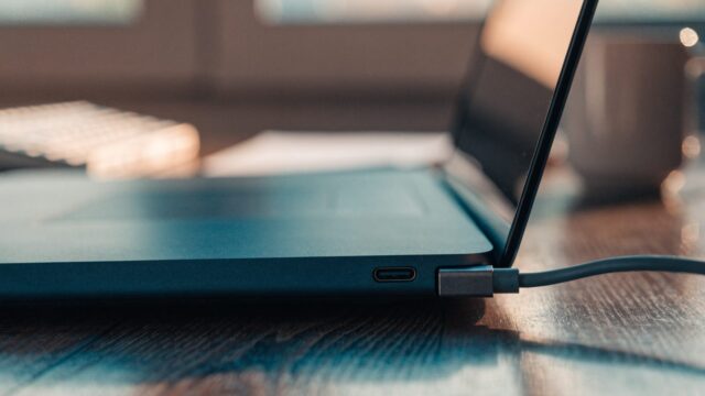 Close-up of a USB-C charger cable plugged into the side port of a dark blue laptop on an office desk