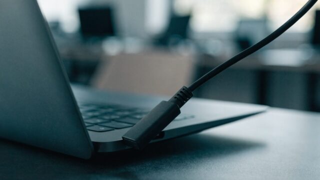 Close-up of a USB-C charger cable plugged into the side port of a graphite black laptop on an office desk