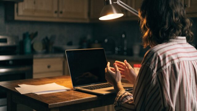 A person seen from behind with hands raised at a matte grey laptop with a dark blank screen on a kitchen table
