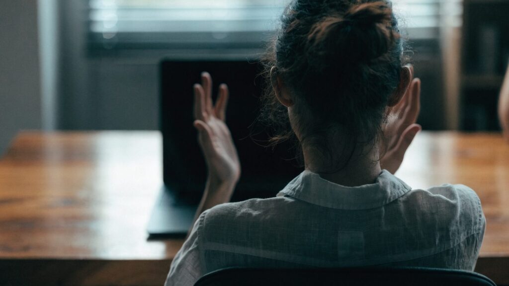 A person seen from behind with hands raised at a graphite black laptop with a dark blank screen on a wooden de