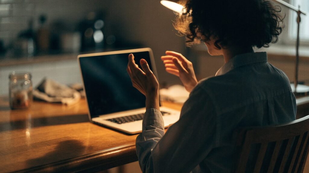 A person seen from behind with hands raised at a silver laptop with a dark blank screen on a kitchen table