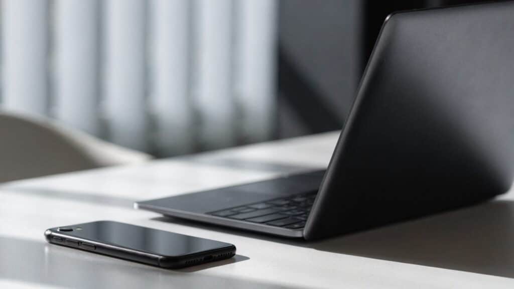 A smartphone lying beside a graphite black laptop on a white desk