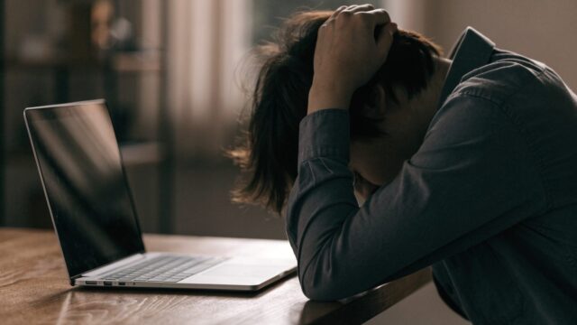 A person resting their forehead on one hand in front of a silver laptop on a wooden desk