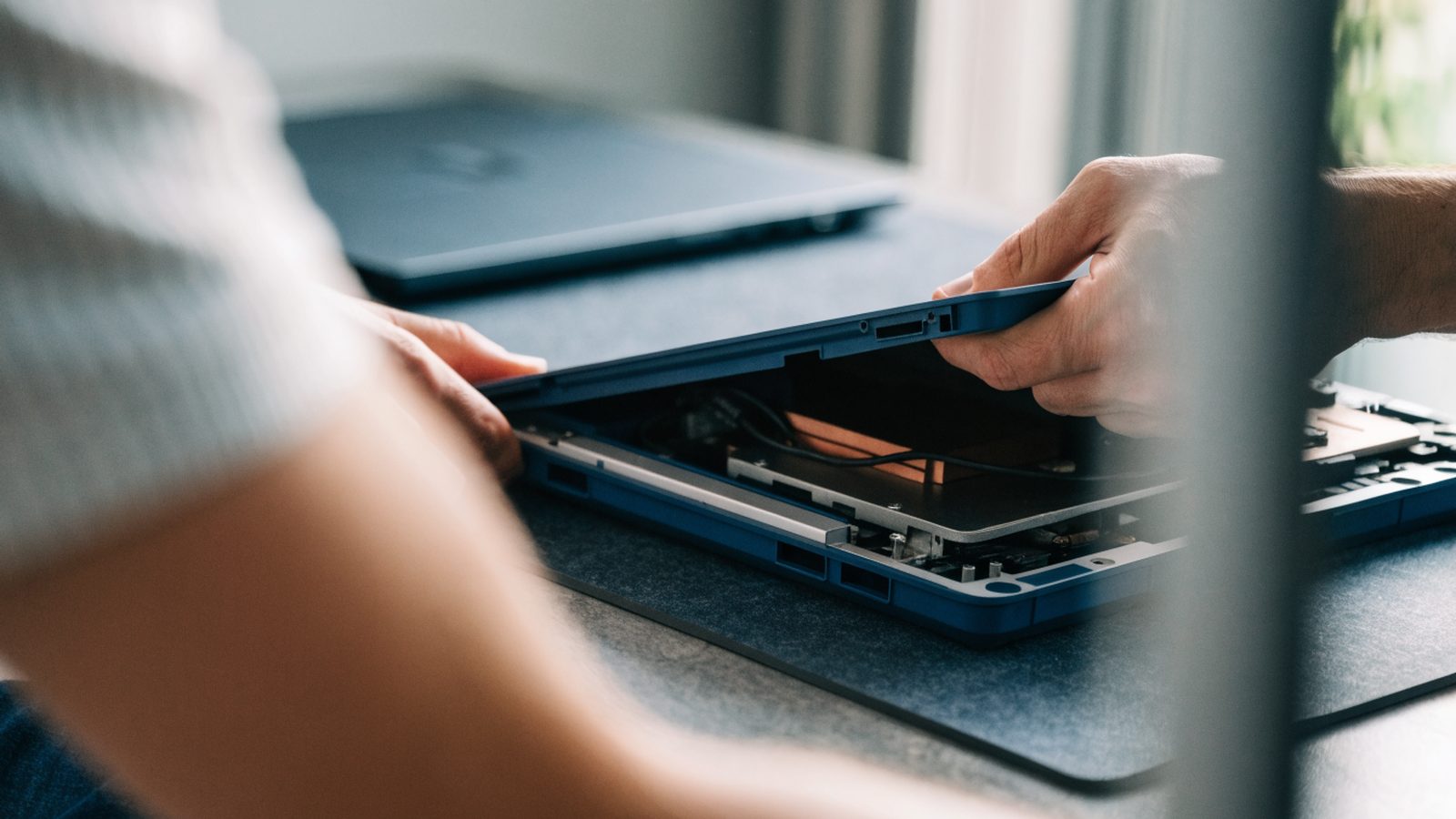 A technician lifting the bottom cover off a dark blue laptop on an anti-static mat