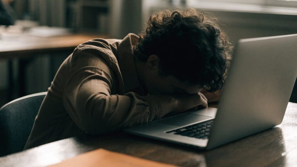 A person resting their forehead on one hand in front of a matte grey laptop on an office desk