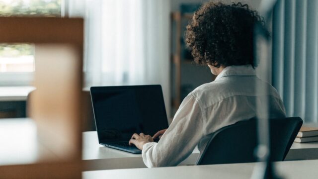 A person seen from behind working on a dark blue laptop in a bright home office