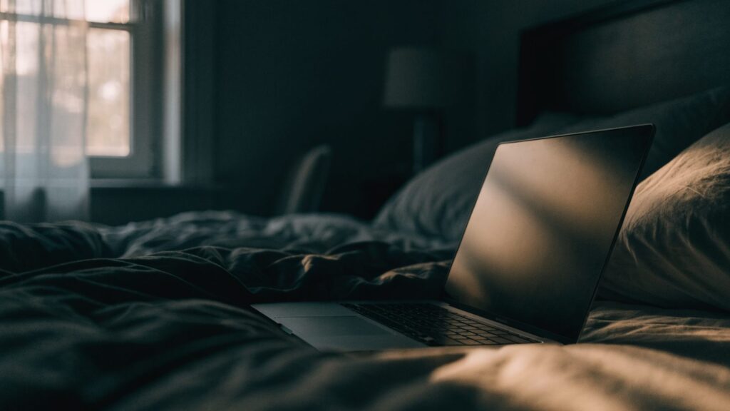 A graphite black laptop glowing softly on a bed in a dark room