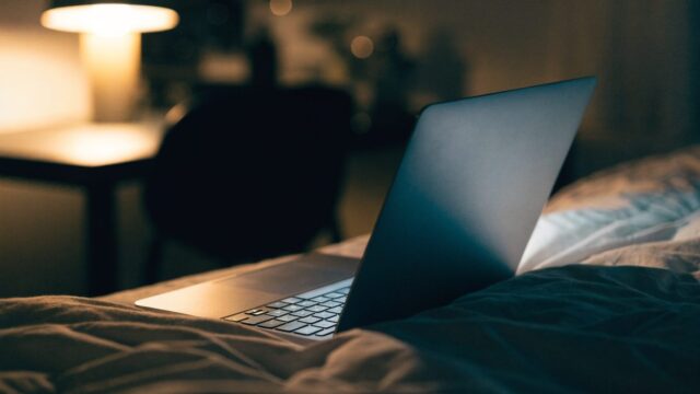 A matte grey laptop glowing softly on a bed in a dark room