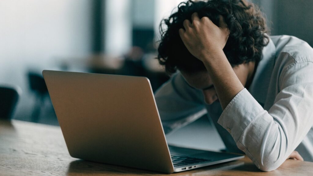 A person resting their forehead on one hand in front of a matte grey laptop on a wooden desk