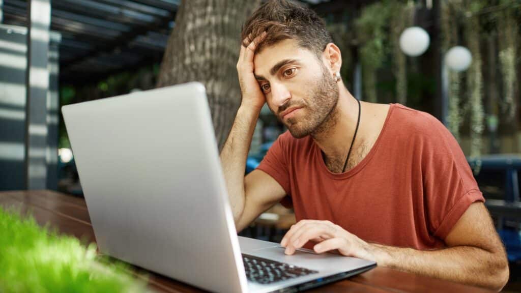 Tired young handsome man sitting with laptop at outdoor cafe, working remote, or studying using park wifi