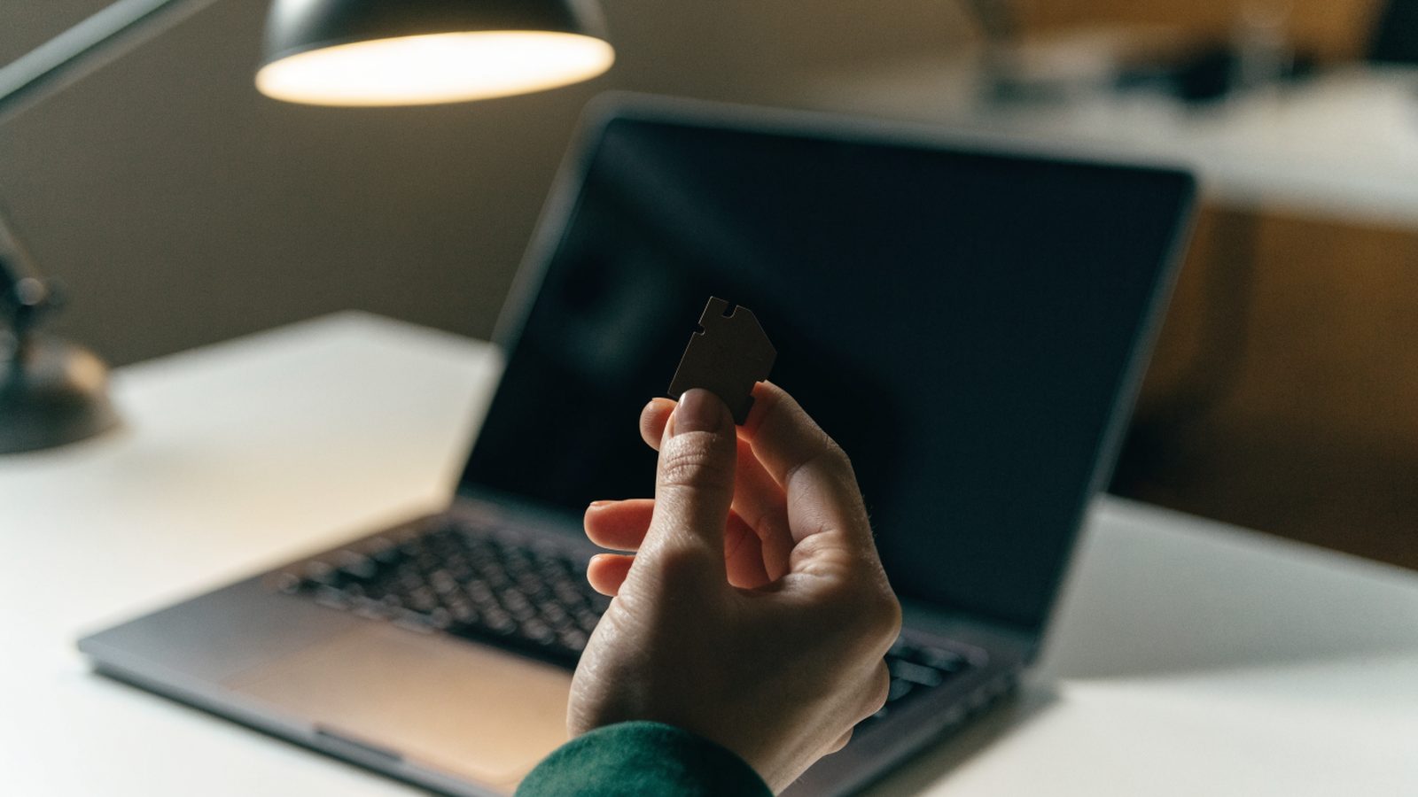 An SD card held between two fingers in front of a matte grey laptop on a white desk
