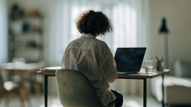 A person seen from behind working on a dark blue laptop in a bright home office