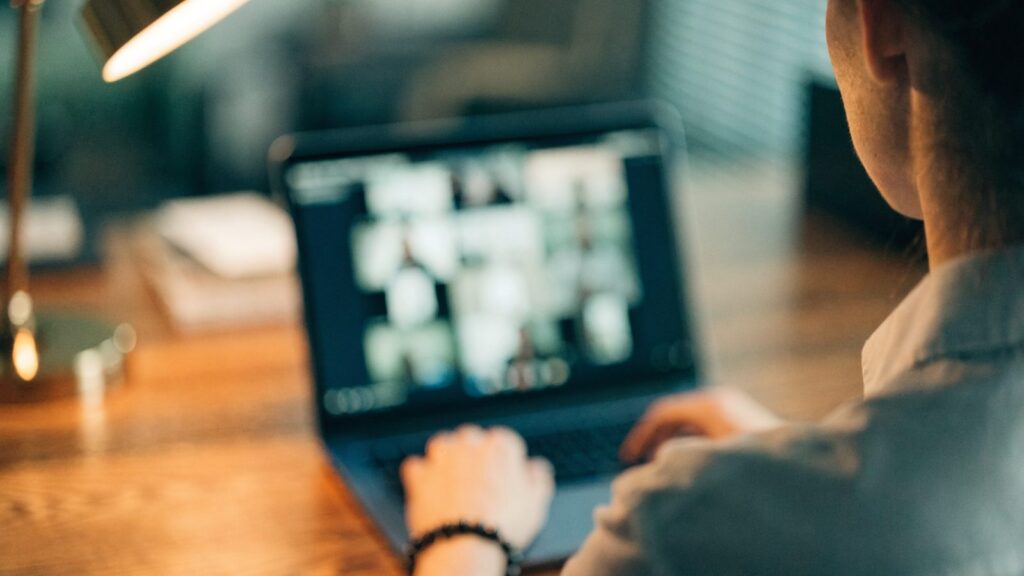 A person seen from behind on a video call on a dark blue laptop on a wooden desk