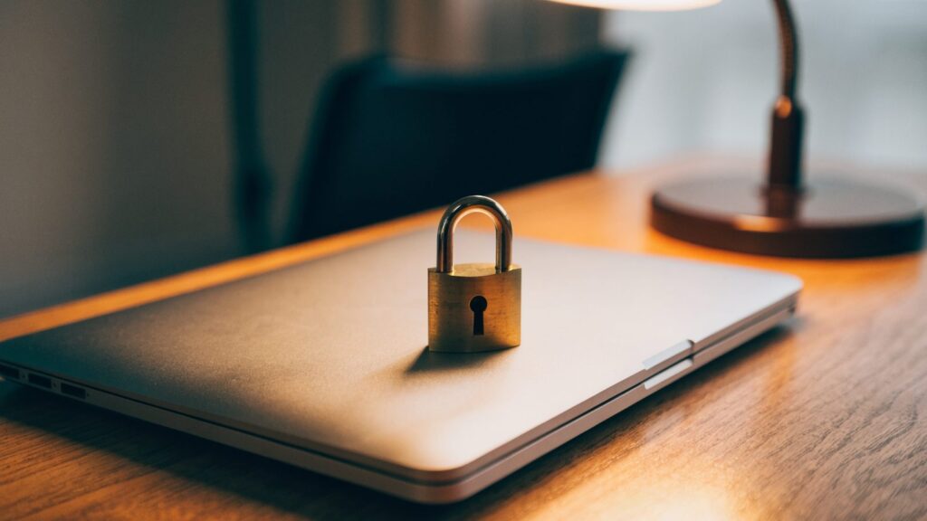 A small brass padlock resting on the closed lid of a silver laptop on a wooden desk