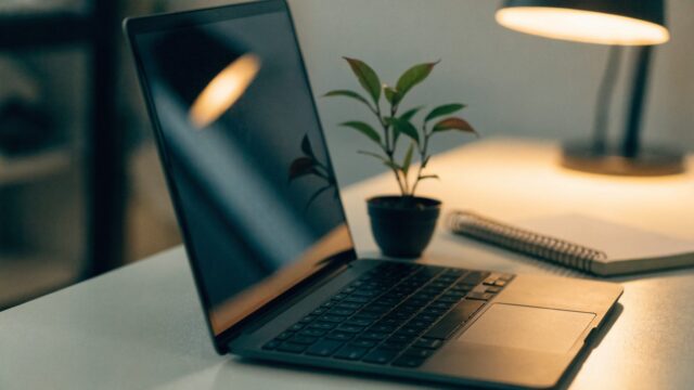 A graphite black laptop open on a white desk with a small plant and notebook beside it