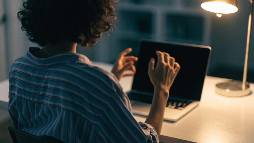 A person seen from behind with hands raised at a silver laptop with a dark blank screen on a white desk