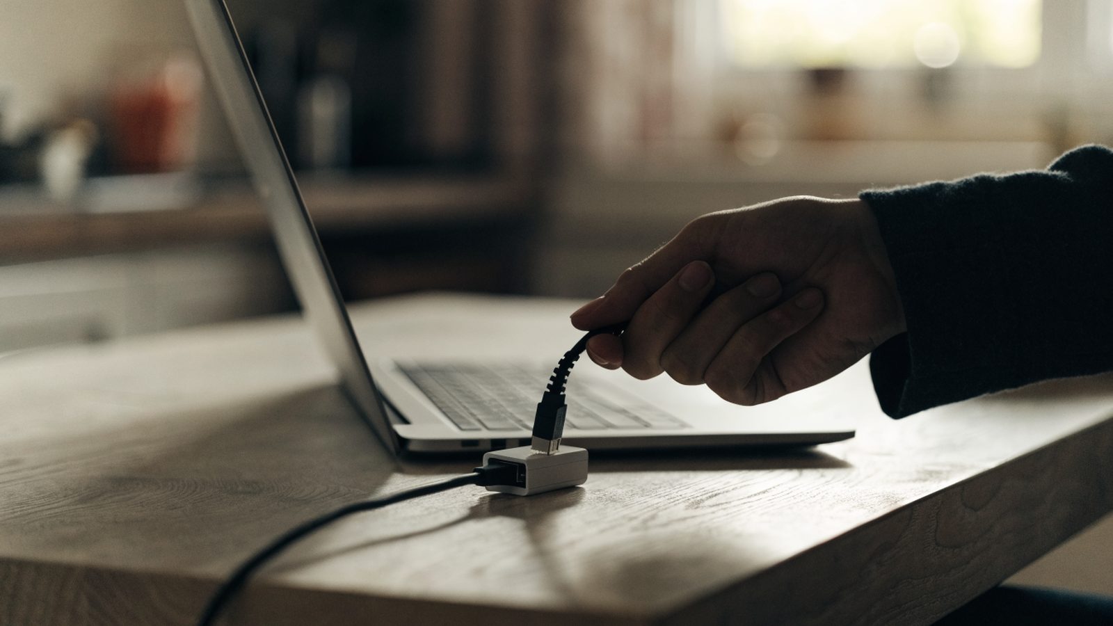 An ethernet cable being plugged into a USB-C adapter connected to a silver laptop on a kitchen table