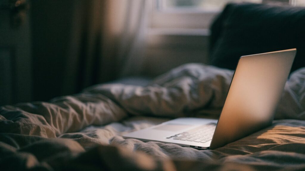 A silver laptop glowing softly on a bed in a dark room