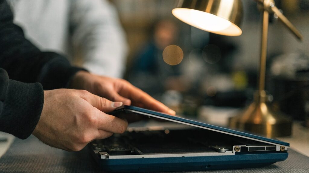 A technician lifting the bottom cover off a dark blue laptop on an anti-static mat