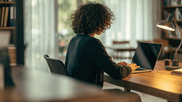 A person seen from behind working on a silver laptop in a bright home office
