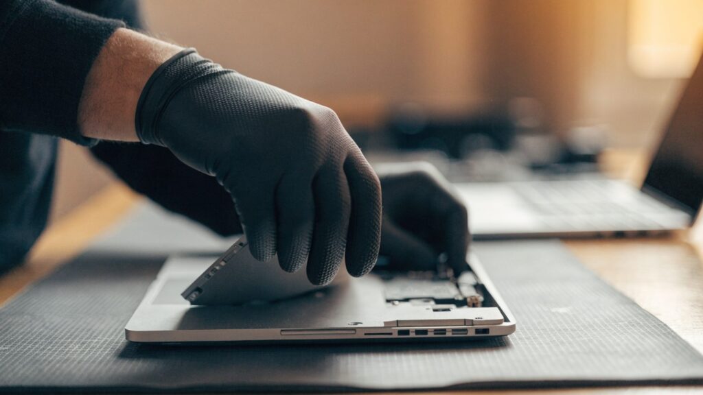 A technician lifting the bottom cover off a silver laptop on an anti-static mat