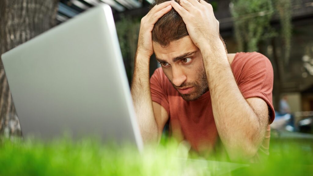 Upset and disappointed handsome guy looking sad at laptop screen while sit outdoors, working on remote…