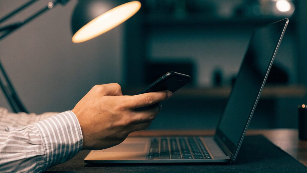 A hand holding a smartphone in front of a matte grey laptop on a dark desk mat
