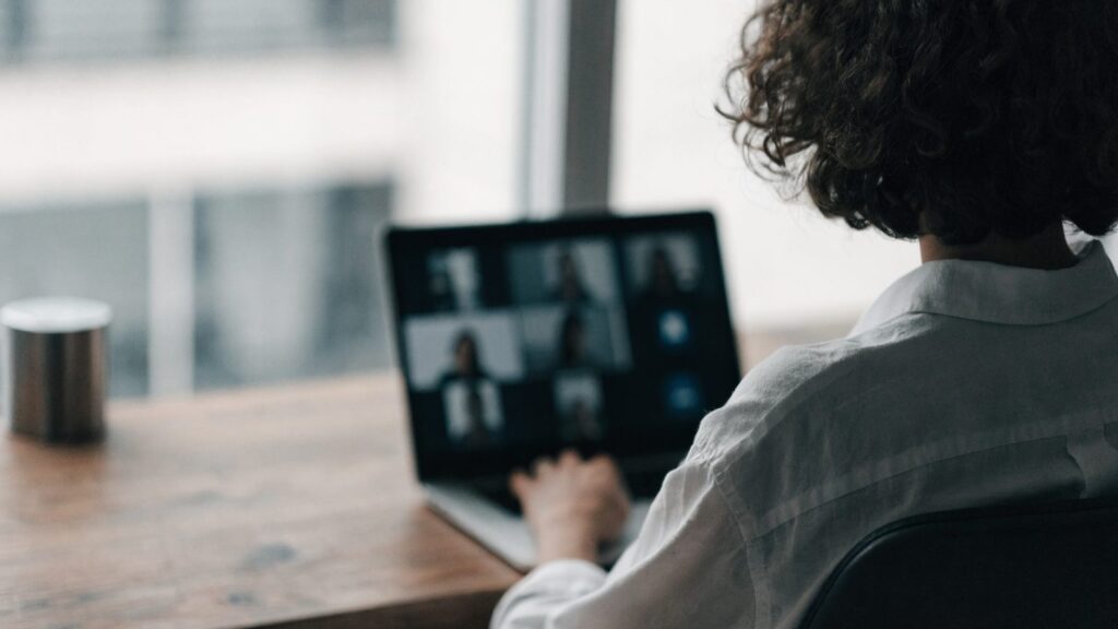 A person seen from behind on a video call on a silver laptop on a wooden desk