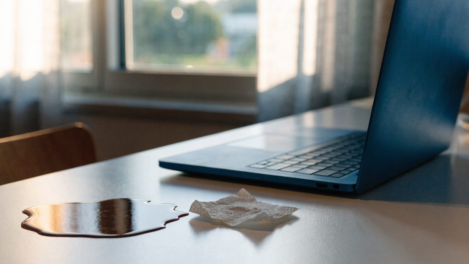 A small puddle of spilled coffee next to a dark blue laptop on a white desk