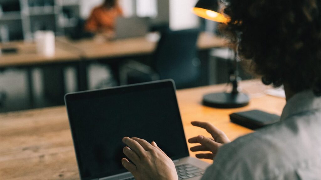 A person seen from behind with hands raised at a matte grey laptop with a dark blank screen on an office desk
