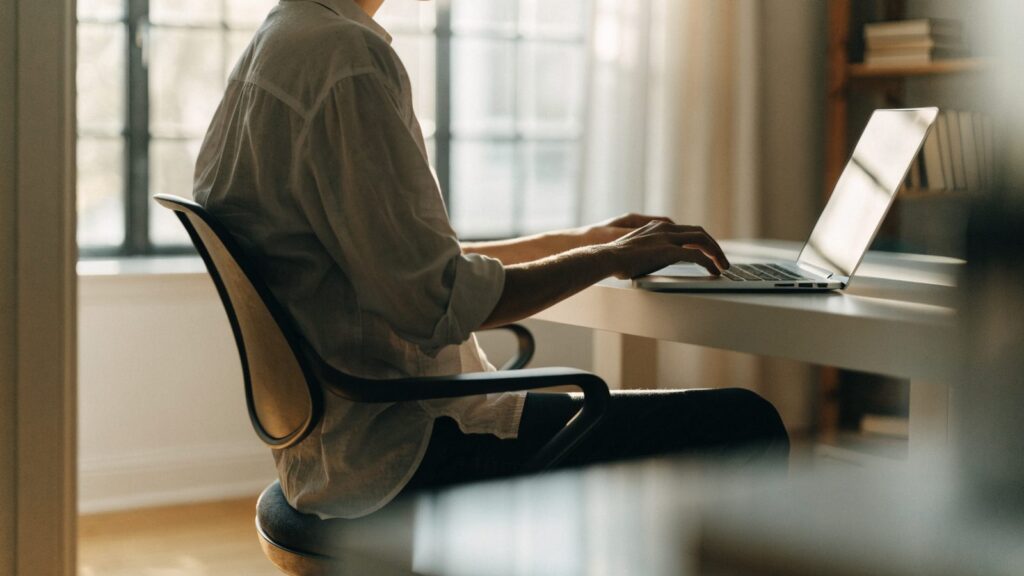 A person seen from behind working on a silver laptop in a bright home office